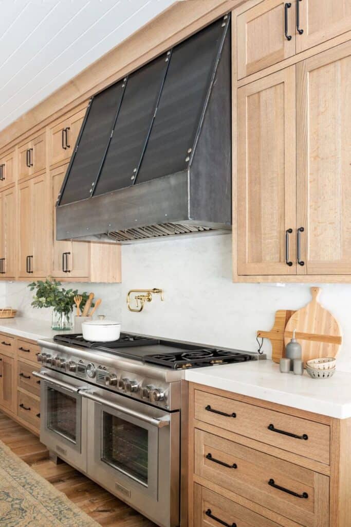 Light wood cabinets with black hardware, white counters and a white backsplash with a very wide oven and black vent hood.