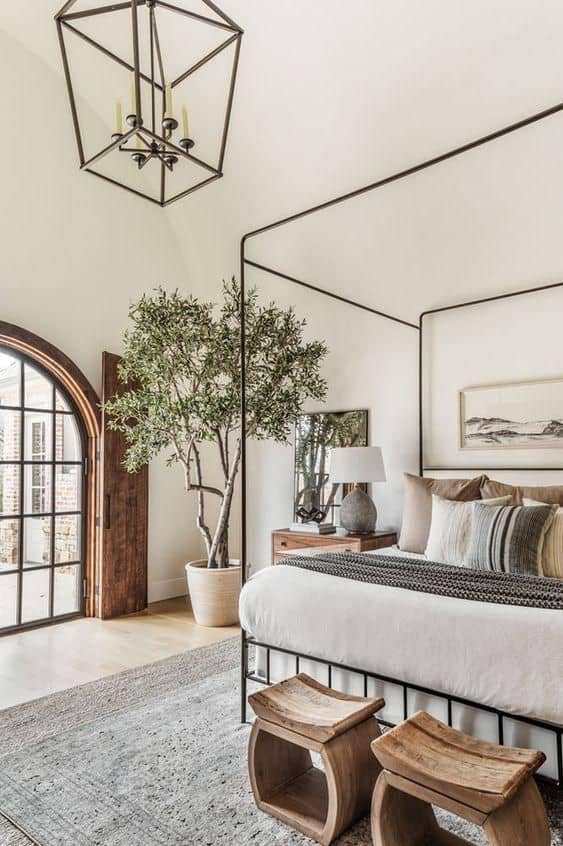 A bedroom with a large arched window, Oyster White painted walls and ceiling, a metal four poster bed and a large olive tree in the corner.