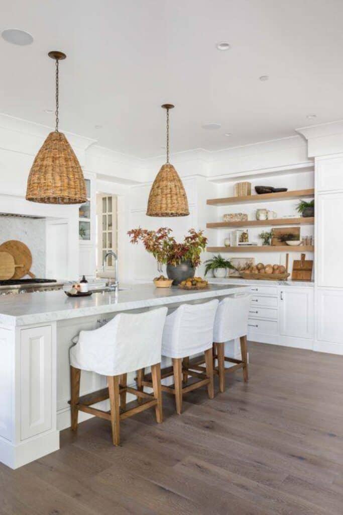 A white kitchen with woven pendents over the island, wood floating shelves and wood cutting boards leaning against the backsplash behind the range.