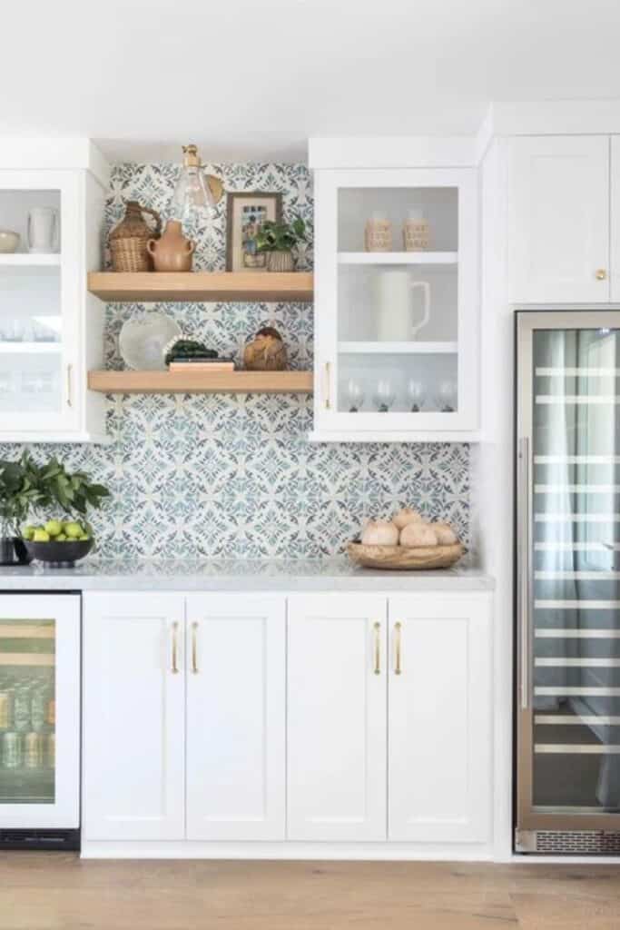 A white cabinet kitchen with a pattered tile backsplash, two wood floating shelves with gold hardware.