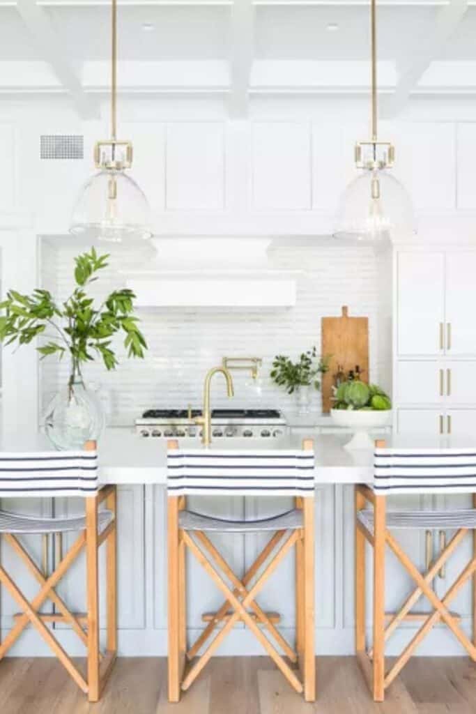 White kitchen with gold hardware, a white tile backsplash, bar stools with striped fabric, and glass pendents over the island.