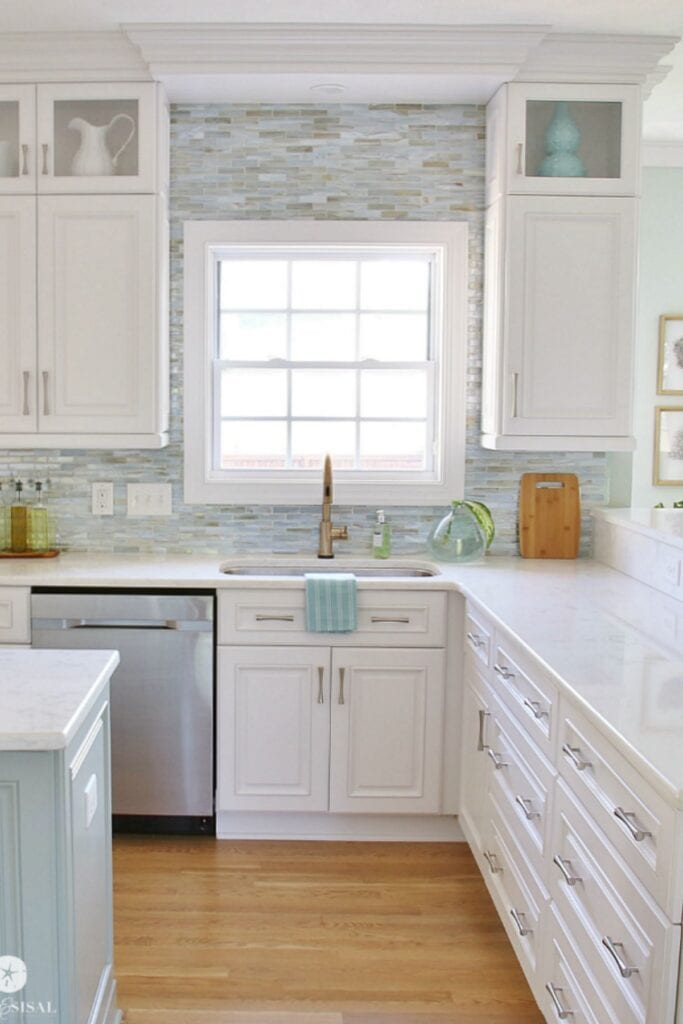 A white kitchen with white cabinets, white counters, silver hardwarer and a large window over the sink.