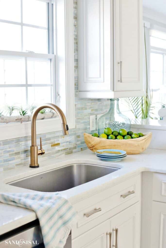 White kitchen cabinets with a blue tile backsplash, gold faucet and silver hardware with a whtie bowl with limes on the counter, a window above the faucet and light blue plates and a striped towel.