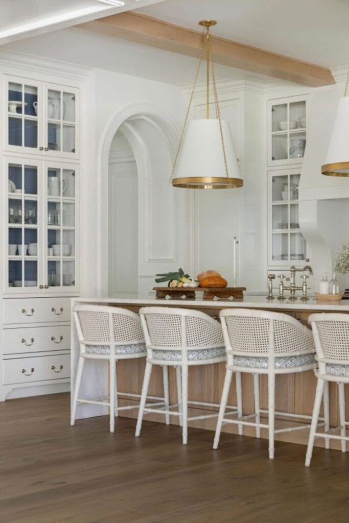 White perimeter kitchens with a wood island, white counters, white pendents with gold trim, glass front cabinets and wicker bar stools.