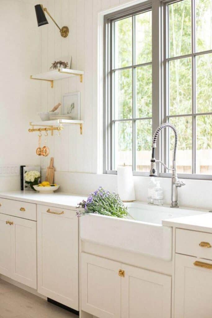 White cabinets with white counters with a farmhouse sink and purple flowers in it, gold hardware with windows above the sink.