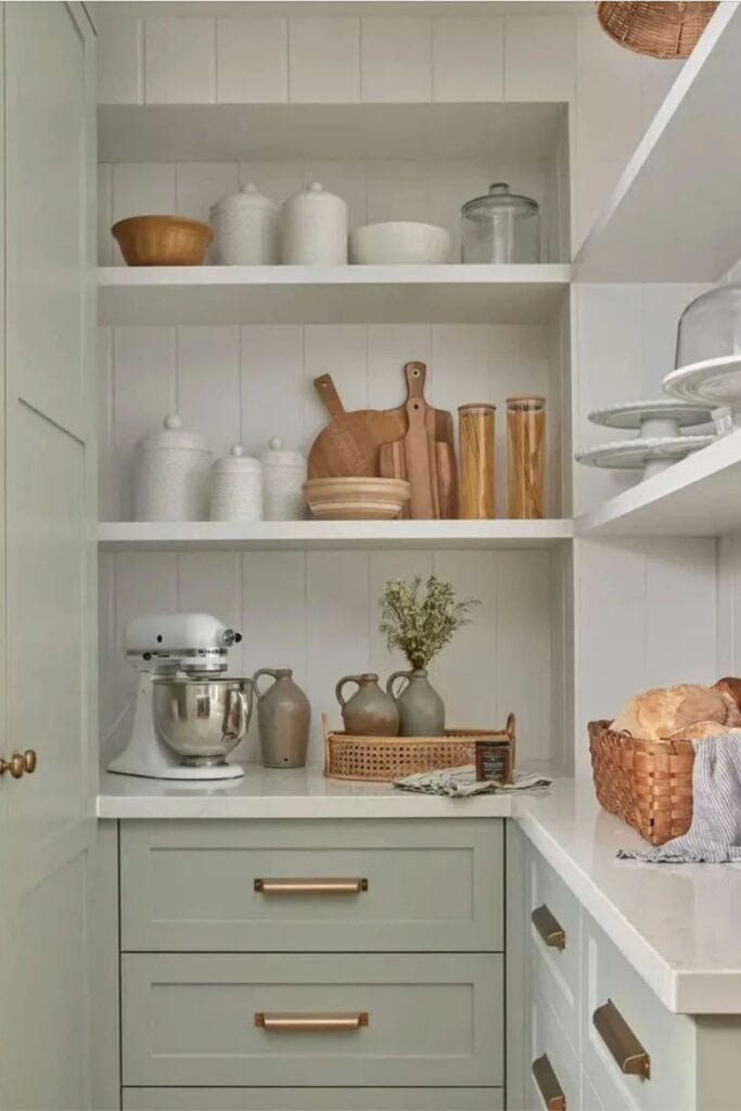 Light green cabinets with whtie counters and white shelves above with white cake plates, wood cutting boards, and a kitchenaid stand mixer.