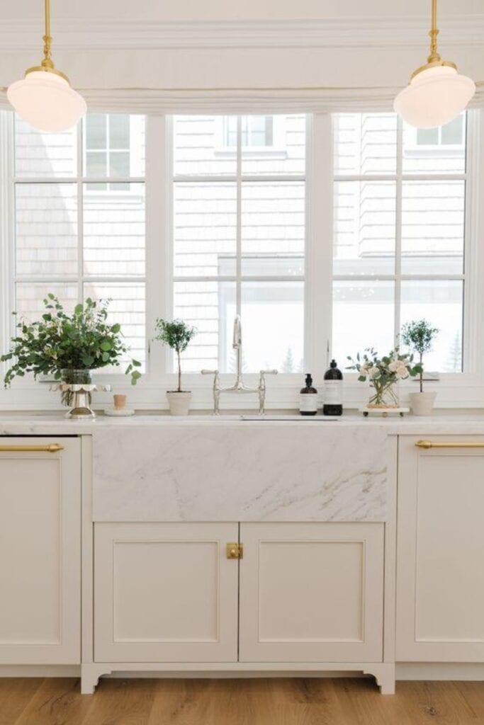 Cream cabinets with a marble farmhouse sink, gold hardware with a silver faucet and large windows above the sink.
