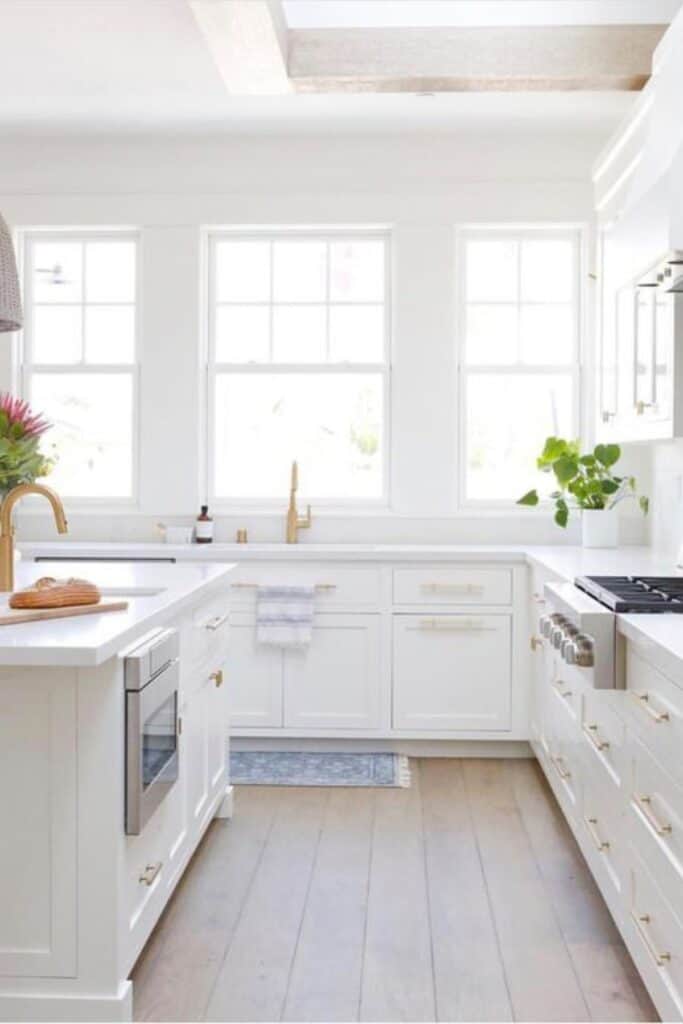 A kitchen with white cabinets, white countertops, tons of windows and gold hardware.