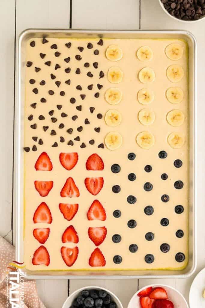 Sheet pan pancakes four ways with one part being sliced bananas, one part being sliced strawberries, one part with blueberries, and one part with chocolate chips.