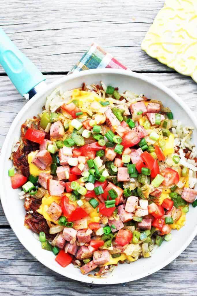 hashbrowns with eggs, tomatoes, ham, green onions, and cheese on a white plate on a wood surface.