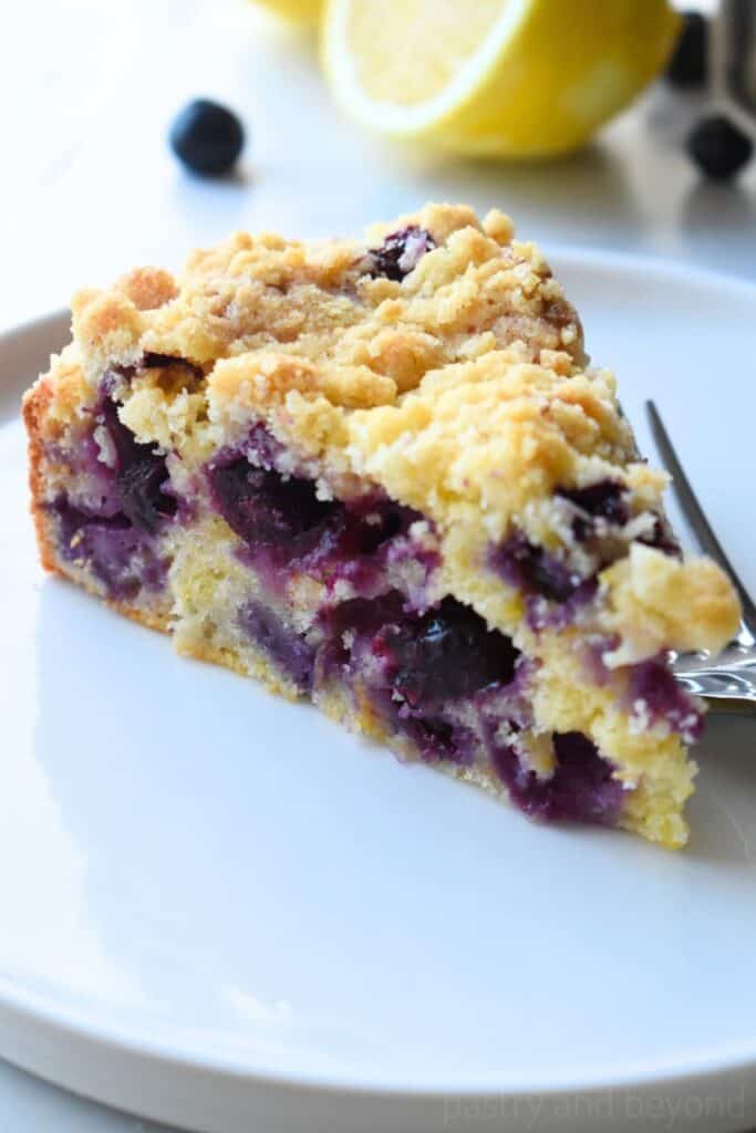 A pie shaped slice of lemon blueberry coffee cake on a white plate and fork.