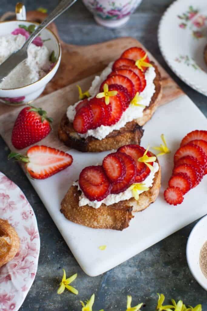 Cinnamon Toast with ricotta cheese and sliced strawberries with yellow flowers on a cutting board that is half wood and half white.