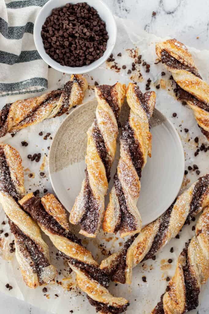chocolate twist pastries on a beige plate with sprinkles of chocolate chips all around and a bowl of them next to a gray and white stripped towel.