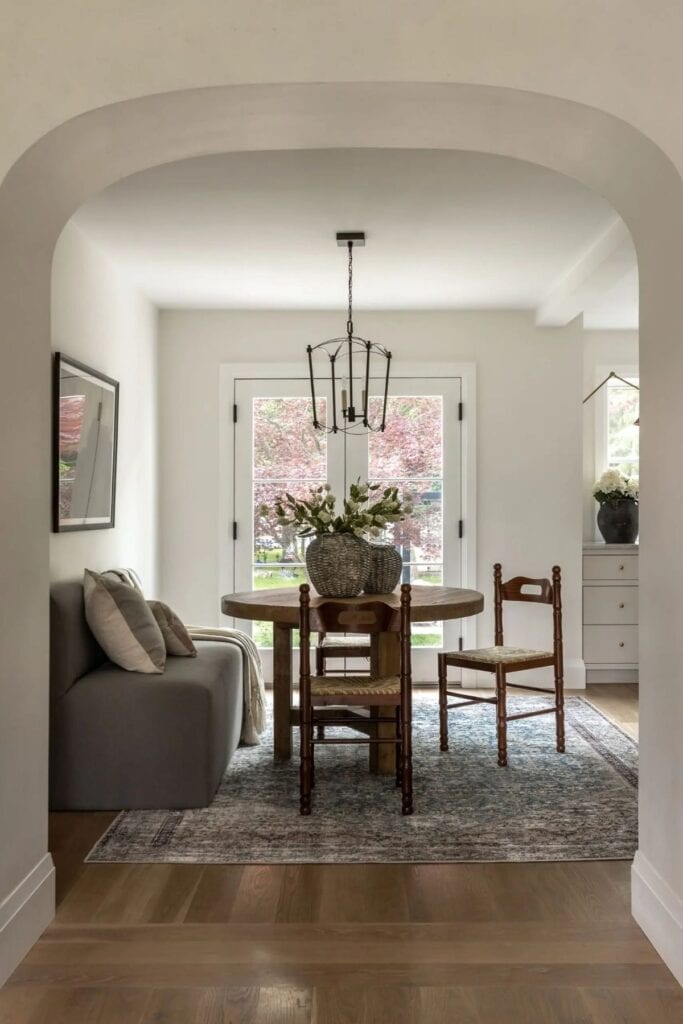 A dining room with a round table and a large vase of flowers with a hanging lantern and the walls are painted Sherwin Williams Oyster White.