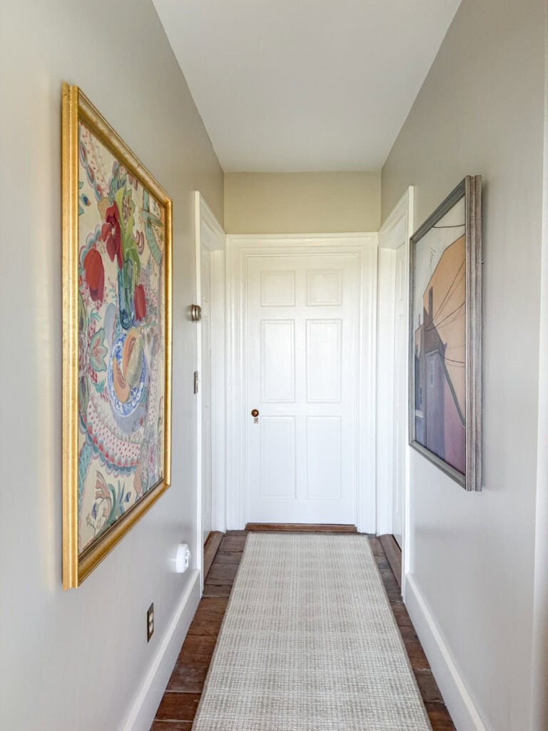 A hallway painted in Ballet White, wood floors iwth a runner rug and large artwork on the walls.