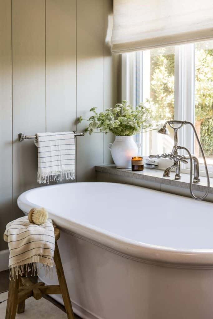 A bathroom with a free standing tub under a large window with vertical plank wall in light gray with silver hardware.