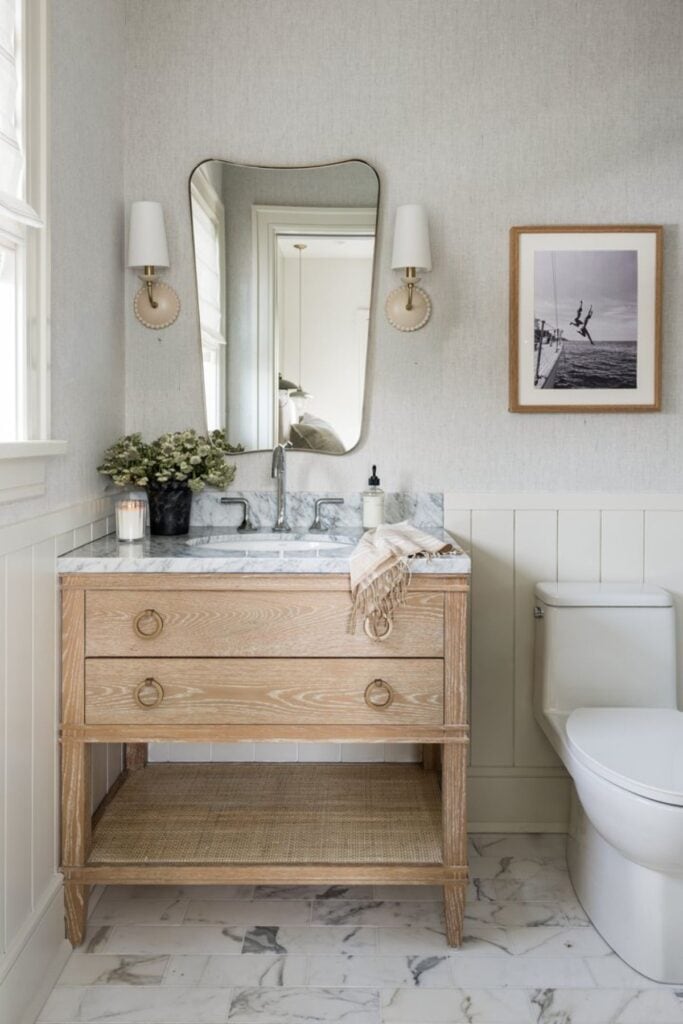 A bathroom with warm whtie walls, a light wood vanity with a marble counter and a black and white photo above the toilet of two people jumping off a boat into water.