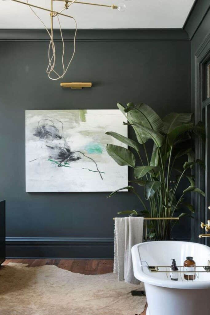 A dark green bathroom that looks almost black with dark wood floors and a huge plant in the corner next to the free standing tub.