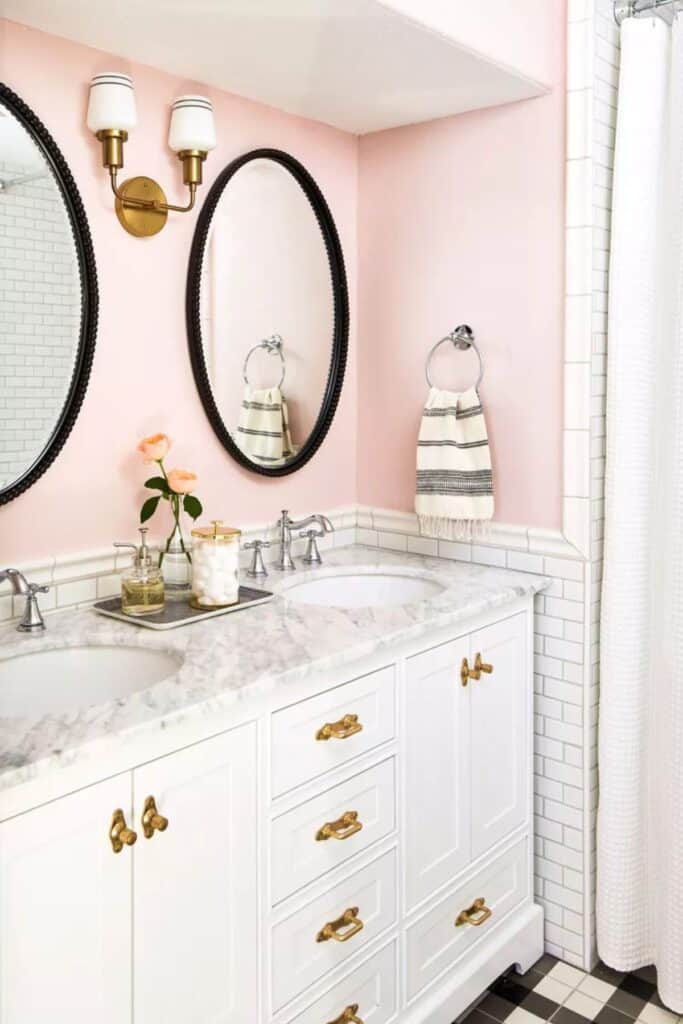 A bathroom with blush pink walls, white tile, white cabinets, marble counters, and black oval mirrors.