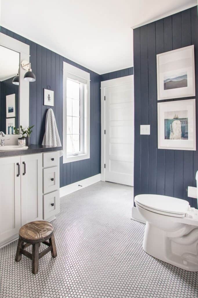 A bathroom with vertical shiplap paianted navy blue, white penny tile floor with nautical prints on the wall.