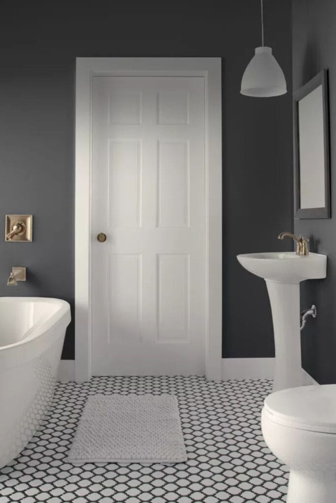 A dark gray bathroom with white fixtures, a white door, and white tile with black grout on the floor.