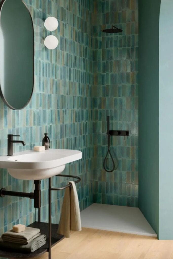A bathroom with teal mosaic tile on the walls, light wood floors and an exposed sink with an oval black rimmed mirror above.