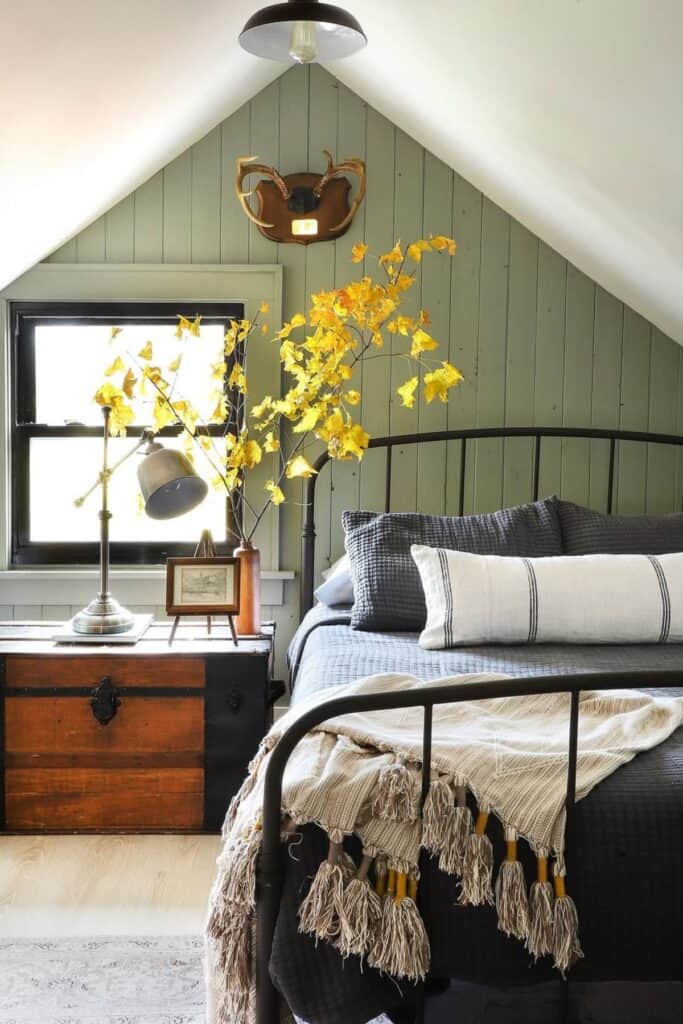A bedroom with a metal bed and dark gray bedding and vertical shiplap painted sage green. the night stand is a chest that is wood and black.
