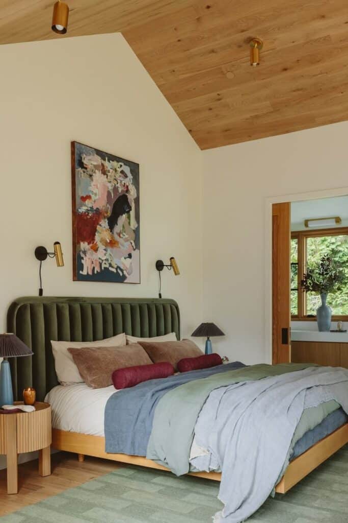 A bedroom with a wood vaulted ceiling, a dark sage green upholstered headboard and blue and sage green bedding.