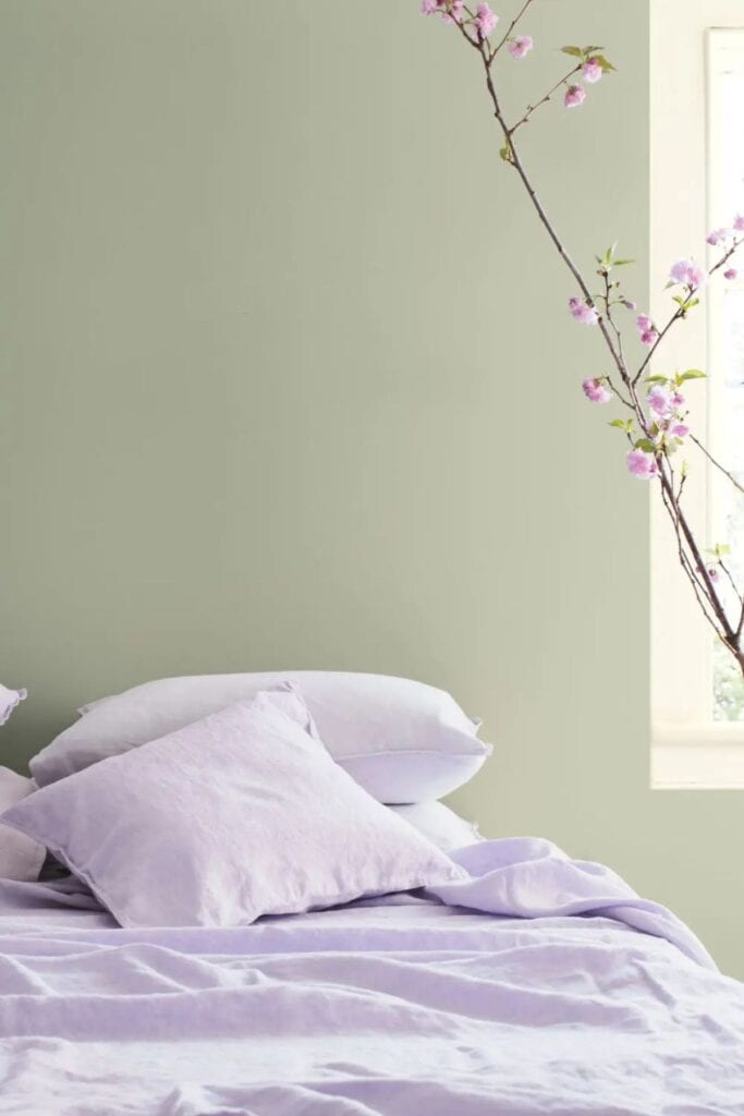 A bedroom with the wall sae green and the bedding lavender and there are lavender flowers.