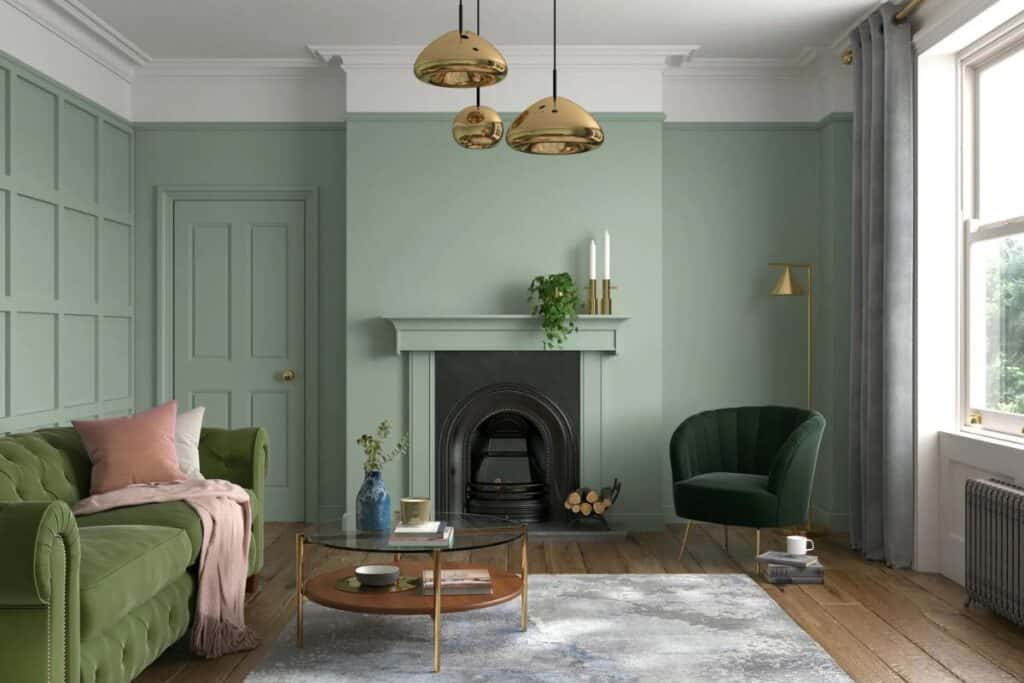 A living room with sage green walls, brass light fixtures, a darker green couch and a forrest green chaiar next to a fireplace.