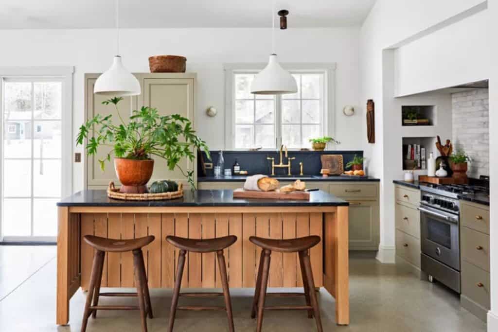 A kitchen with sage green perimeter cabinets and a wood island and they all have black counters with white walls.