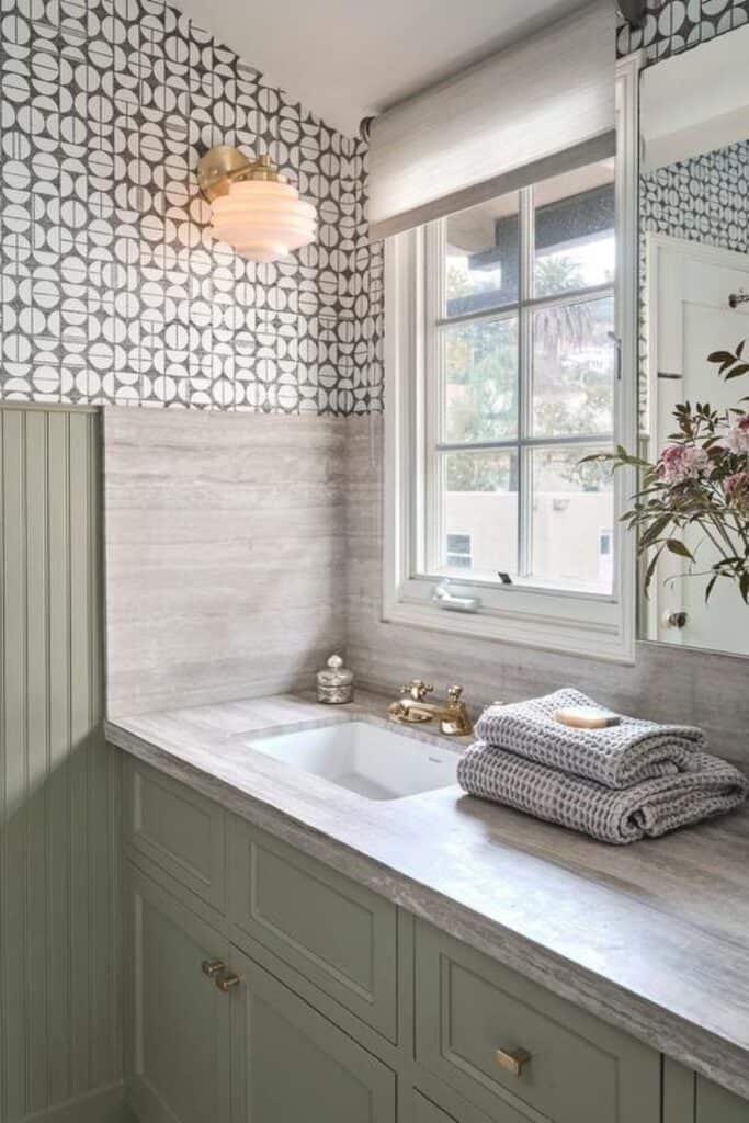 A bathroom with beadboard and a vanity painted sage green and the countertops are gray and there is a geometric wallpaper with a window above the sink.