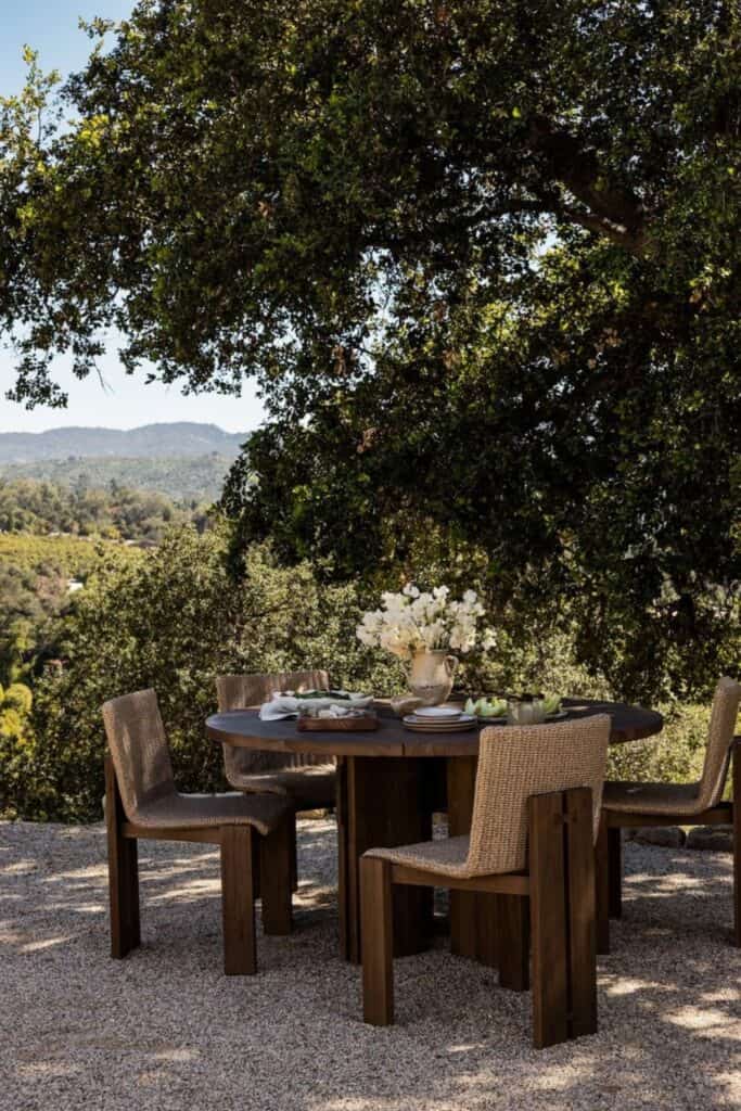 A round table and chairs sitting in gravel with the view of a mountain range and lots of trees.