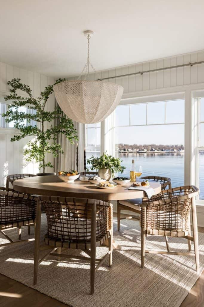 A round table with a large woven chandelier, a tall tree in the corner and a wall of windows with the view of a lake.