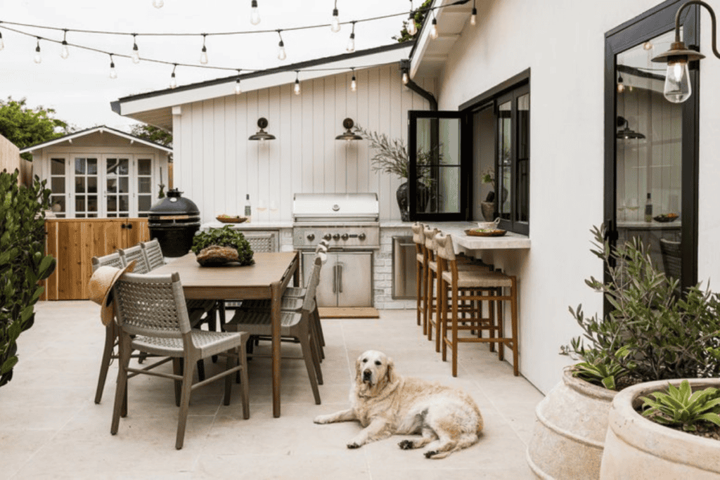 An outdoor kitchen and dining area with string lights, a rectangular table and chairs, a counter with bar stools and a large grill, and a dog laying on the patio.