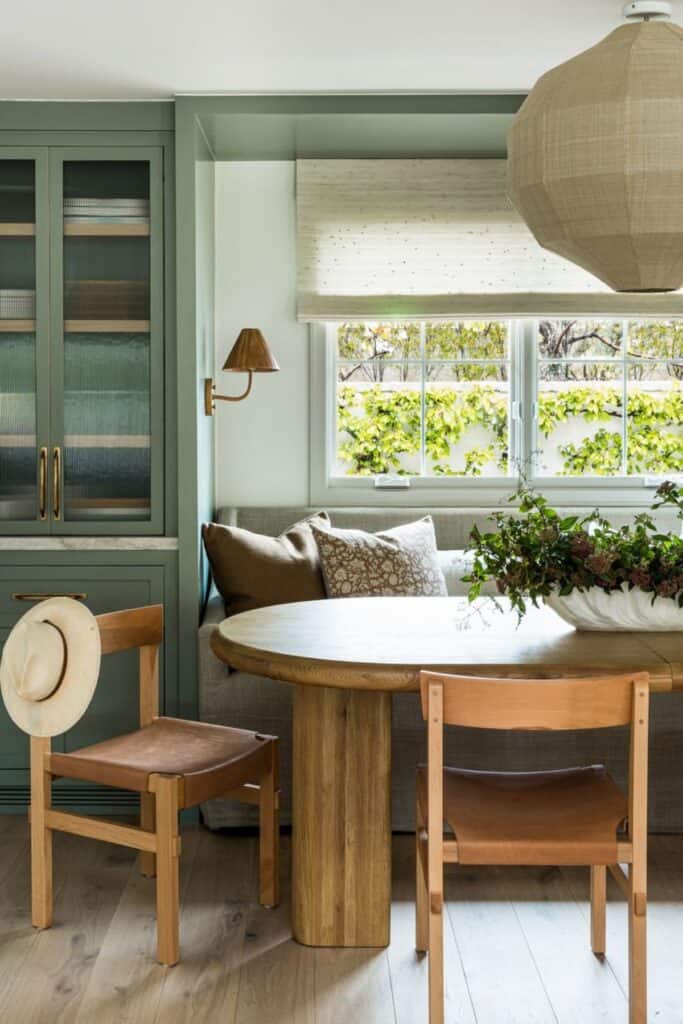 An oval dining table with a bench on one side with windows above and a large round light fixture.