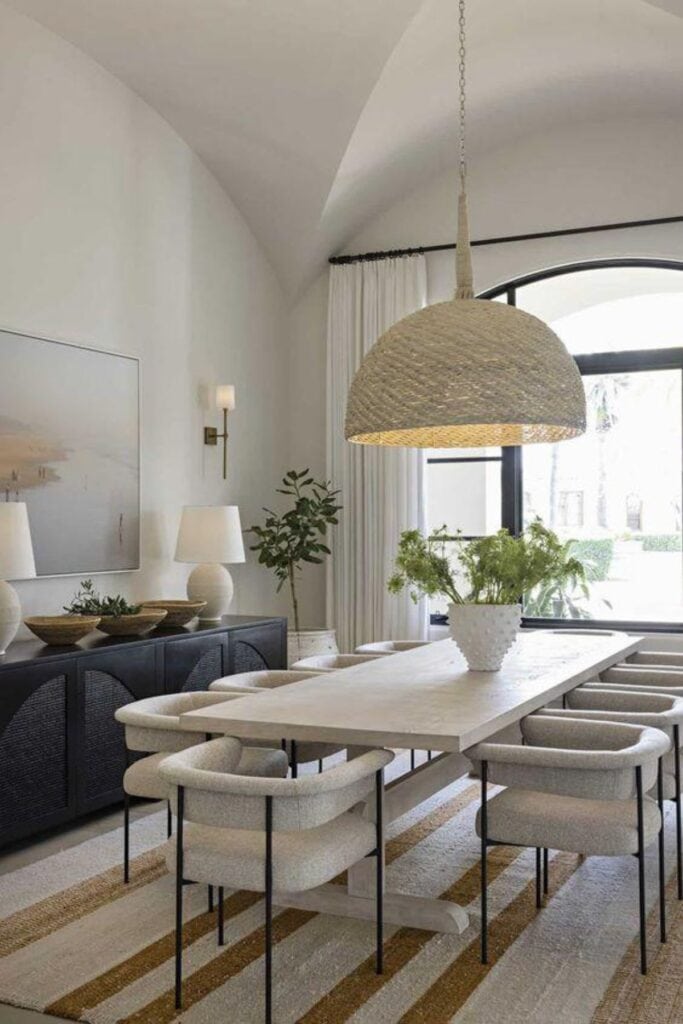 A dining room with a white wood table, curved chairs, a black sideboard with two lamps and three bowls on it with a large window at one end of the room.