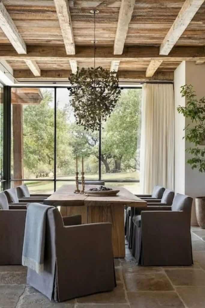 A rustic wood dining table with gray upholstered chairs and a huge window at the end with white curtains.