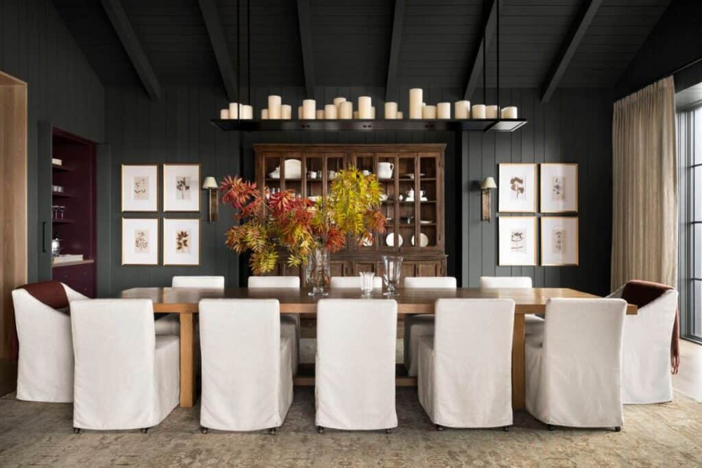 A ver long table and chairs that have white covers, vertical shiplap walls painted a dark color and a wood hutch.
