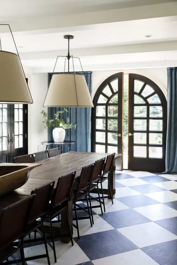 A dining room with a long wood table, two large pendents, arched french doors with tall blue curtains.
