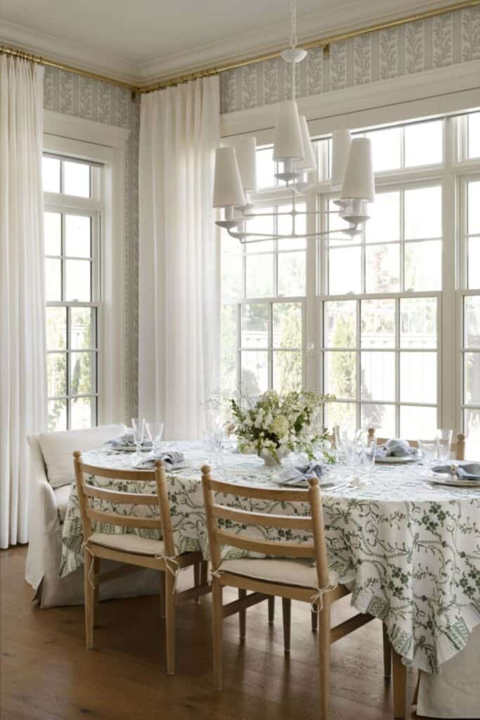 A dining table with a tablecloth on it that is white with green flowers, wood chairs on the sides with large white upholstered chairs at the head and lots of windows.