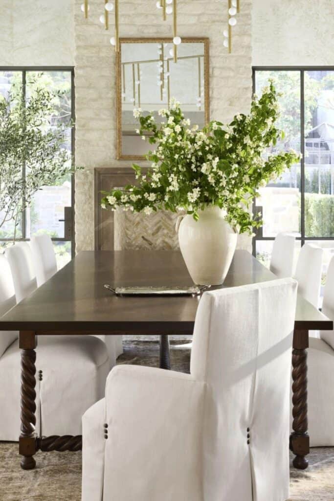 A dining table with curved legs, white upholstered chairs and a large white vase with branches in the vase that have leaves and small white flowers.