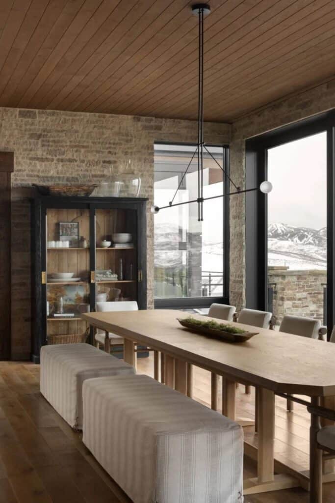 A dining room with a bench on one side of the table and chairs on the other, the walls are stone and there is a large black curio cabinet.