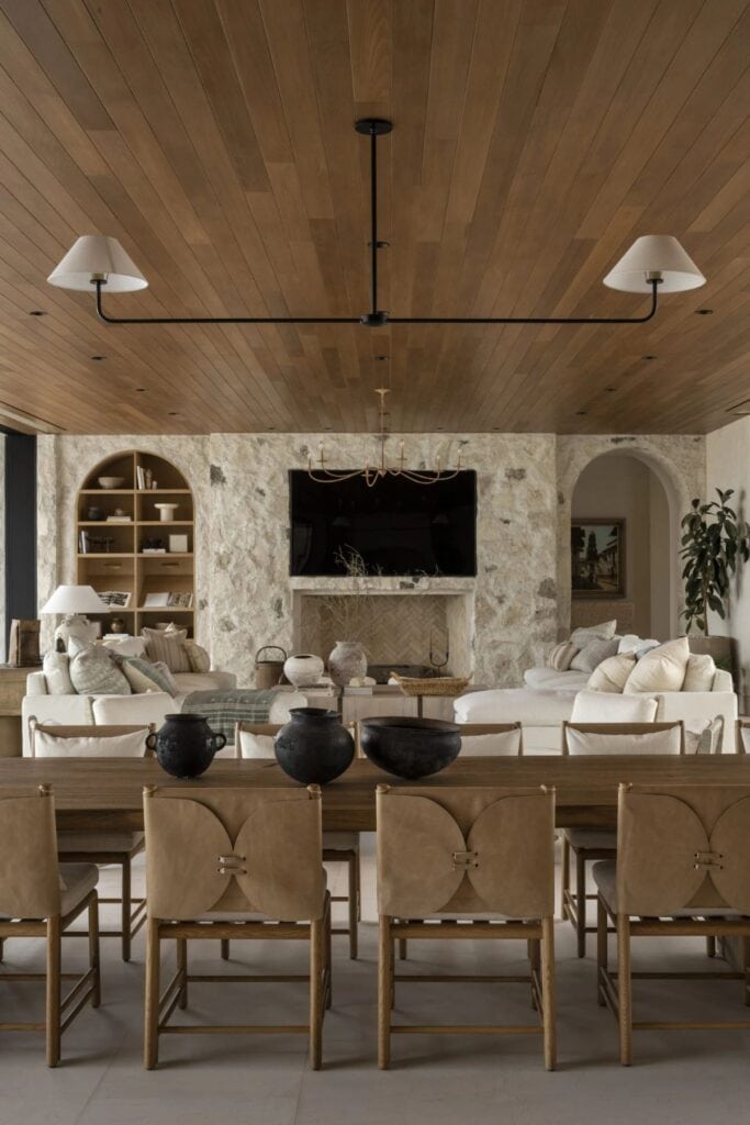 A dining room that looks onto the family room. It has chairs that look like envelopes on the back, a wood ceiling, three black bowls on the table and the fireplace wall is stone.