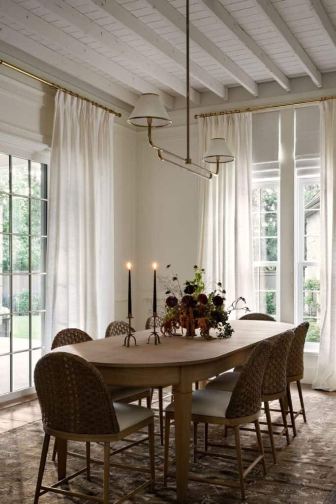 An oval wood dining table with a flower arrangement and two candles, tall windows and tall white curtains.