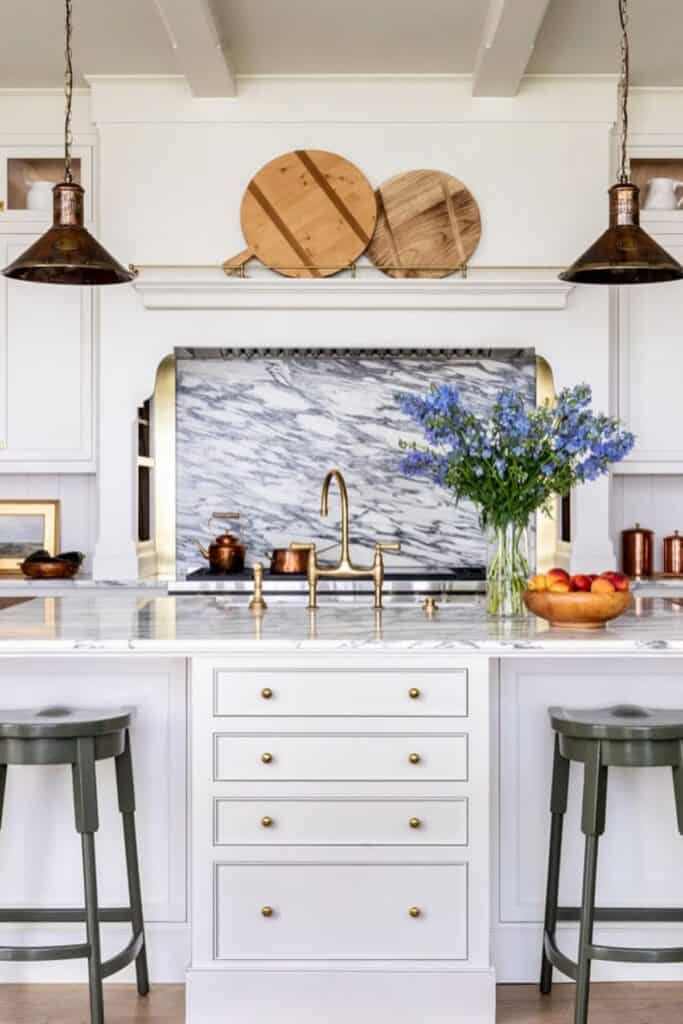 A white kitchen with a marble backsplash and cutting boards above the cabinets.