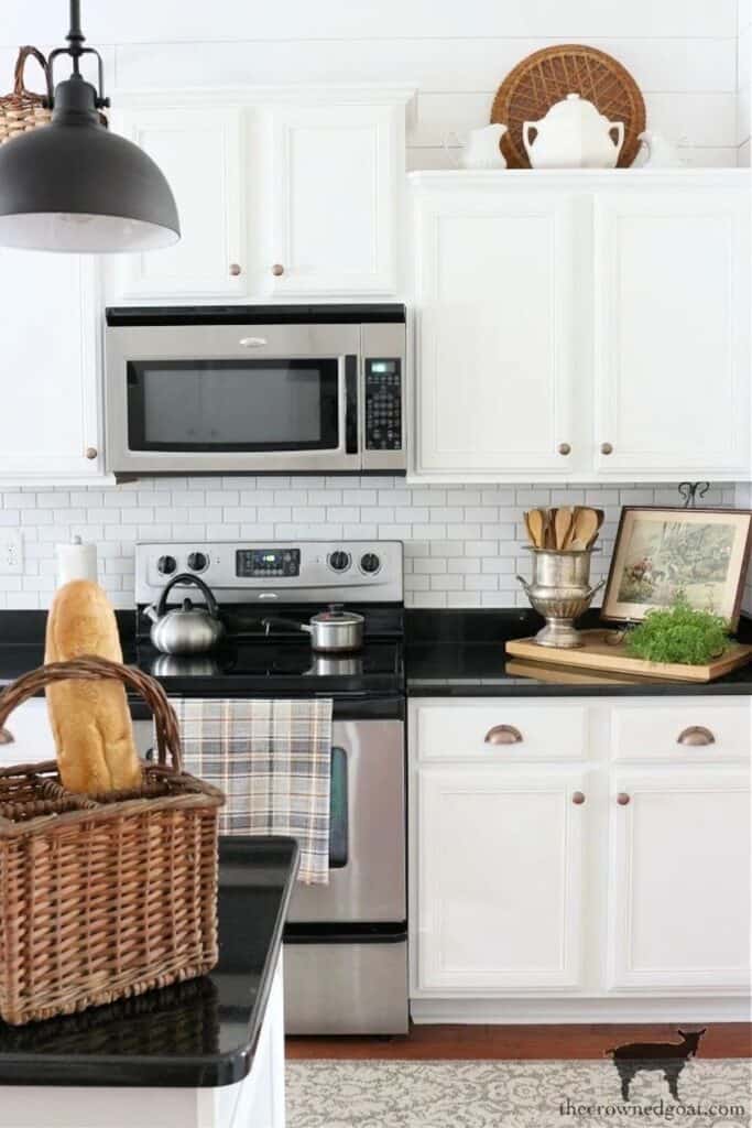 A white ktichen with black counters and above the upper cabinets are white pitchers and behind them a woven basket.