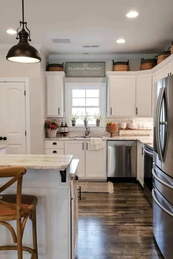 A white kitchen with woven baskets above the upper cabinets and a sign above the sink window that says flower market.