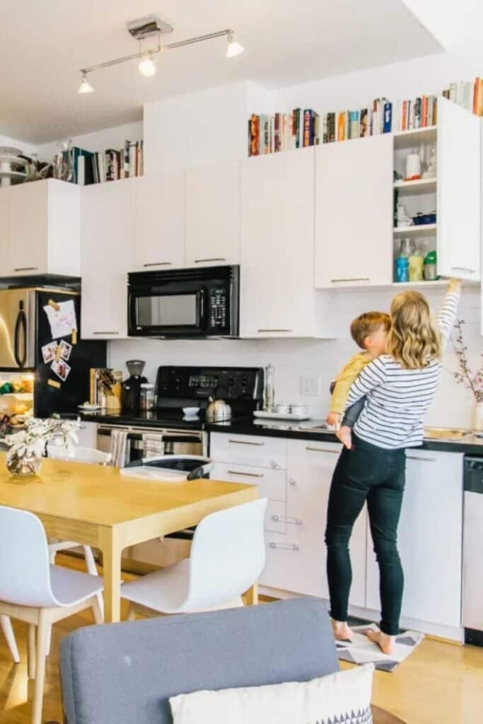 A white kitchen with black and stainless appliances and books above the cabinets with a woman holding a child getting something out of the upper cabinet.