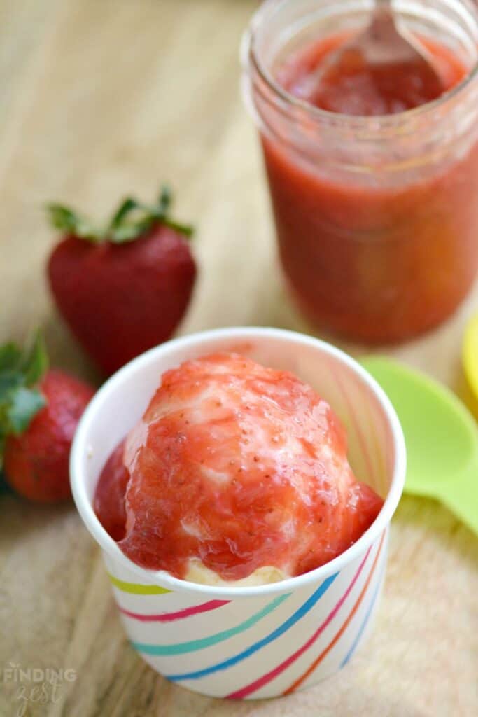 A strawberry rhubarb sauce over ice cream in a paper cut with multi colored lines on it and a lime green plastic spoon next to it sitting on a wood surface.